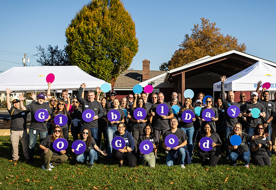 A group of people outdoors hold purple and blue circles spelling "Global Day of Good," standing in front of white tents on a sunny day with green grass and autumn trees.