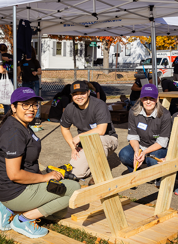 Three people wearing purple hats and black shirts kneel around wooden picnic table parts, assembling them outdoors under a tent. They are smiling, using tools, and working as a team on a sunny day.